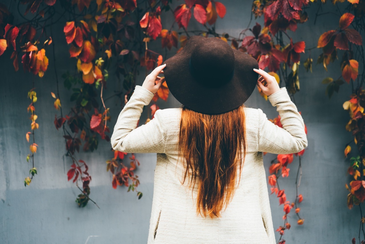 Rückansicht einer schönen jungen Frau mit Hut, die auf einer herbstlichen Straße steht. Trendige junge Frau mit roten Haaren im Herbst im Park.